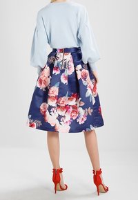 Kvinna iklädd en ljusblå tröja, en marinblå blommig midiklänning och röda högklackade sandaler, stående mot en vit bakgrund.