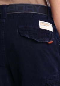 Detailní záběr na tmavě modré cargo kalhoty Superdry s kapsou na zadní straně, na které je bílá a oranžová značka značky a černý pásek s logem Superdry.