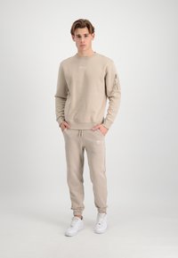 Beige sweatshirt med rund hals og sidelomme med lynlås, parret med matchende beige joggers. Har ribbede manchetter og logodetalje.