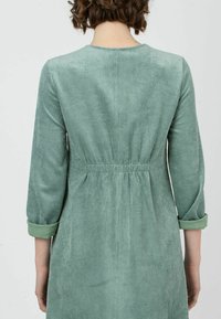 Vestido de pana verde con cintura entallada, mangas tres cuartos, escote redondo y textura fruncida en la parte trasera, mostrando tela acanalada.