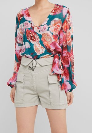 Kvinde iført en teal blomsterwrapbluse med pink og røde roser, kombineret med højtaljede beige shorts med store sidelommer og bælte.