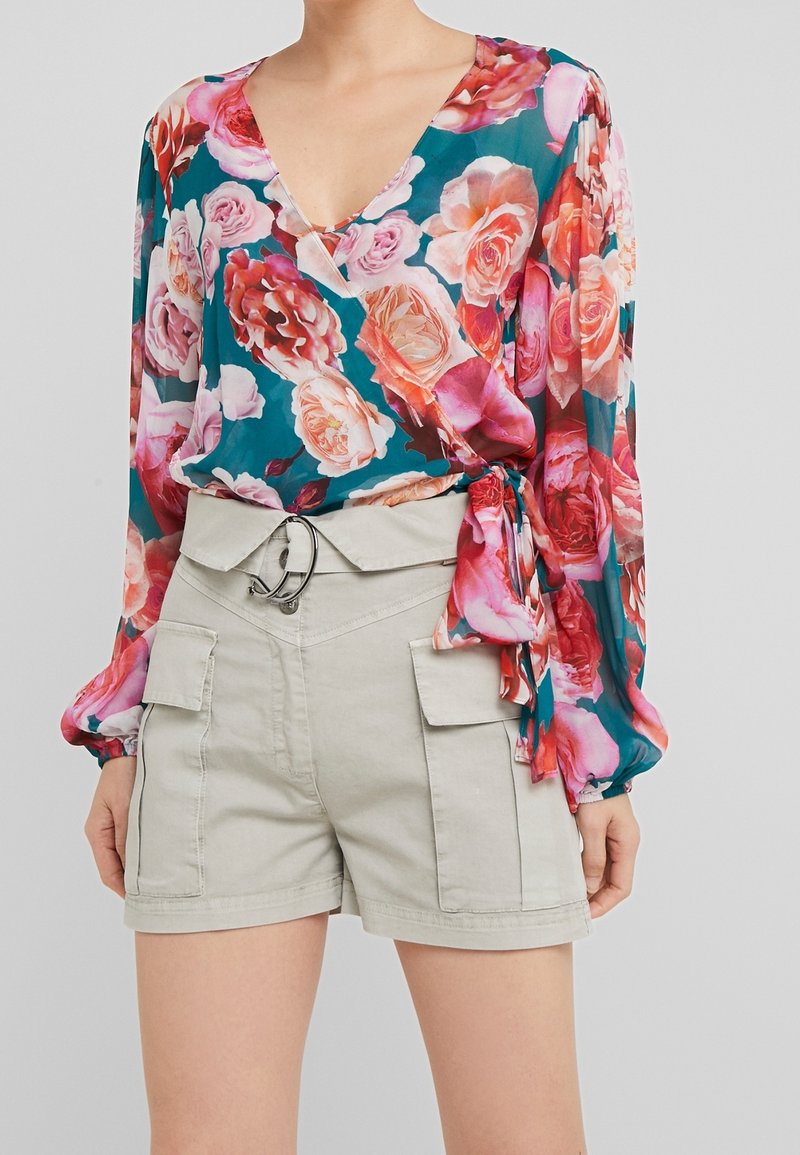 Femme portant un chemisier cache-cœur floral bleu sarcelle avec des roses roses et rouges, associé à un short beige taille haute avec grandes poches latérales et ceinture.