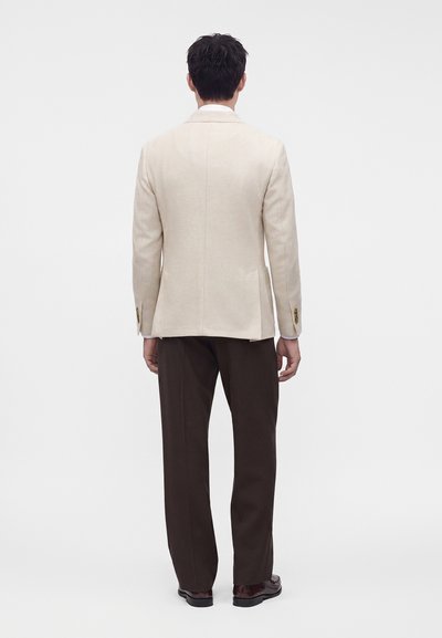 Un homme de dos porte un blazer crème, une chemise blanche, un pantalon marron foncé et des chaussures habillées marron foncé sur un fond blanc uni.