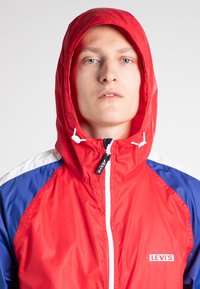 Veste coupe-vent rouge à capuche avec des panneaux bleus et blancs, dotée d'une fermeture éclair à l'avant, de cordons ajustables et d'un logo sur la poitrine.