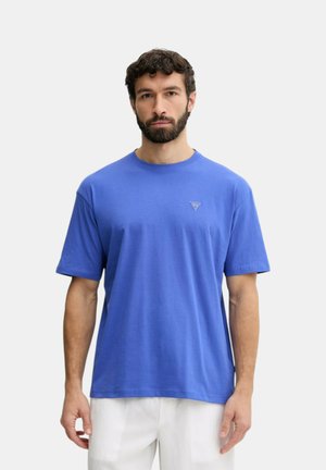 Uomo con capelli corti e ricci e barba, che indossa una maglietta blu semplice a maniche corte e pantaloni bianchi, in piedi davanti a uno sfondo chiaro.