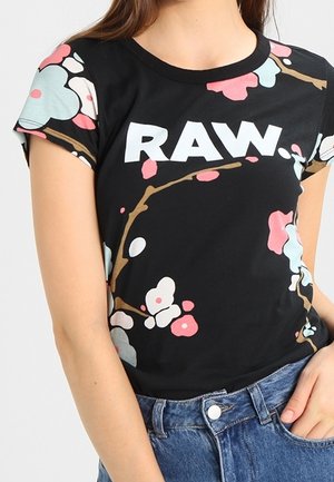 Czarny T-shirt z białym napisem "RAW" oraz pastelowym różowym, niebieskim i białym wzorem kwiatowej gałązki, noszony z niebieskimi dżinsami.