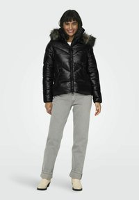 Chaqueta acolchada negra con cierre de cremallera, capucha forrada de piel sintética y patrón de acolchado diagonal. Combinada con pantalones gris claro y botines crema.