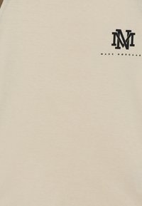 Beige kortärmad t-shirt i mjukt tyg. Har svart logotyp med "M" och "MADS NØRGAARD" på vänster bröst. Slät textur.