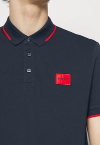 Homme portant un polo bleu marine avec un liseré rouge sur le col et les manches, patch rectangulaire rouge avec "HUGO" sur la poitrine gauche.