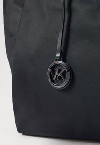 La borsa a tracolla in tessuto nero presenta un grande charm con logo in rilievo in argento metallizzato, con una finitura testurizzata e un design elegante e minimalista.