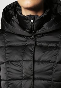 Schwarze gepolsterte Jacke mit Reißverschluss, hohem Kragen und verstellbare Kapuze. Merkmale ein gestepptes Design mit glatter, glänzender Textur.