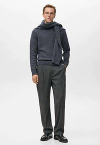 Uomo che indossa un maglione grigio scuro con una sciarpa abbinata drappeggiata sulle spalle, pantaloni grigi e scarpe nere, in piedi davanti a uno sfondo semplice.