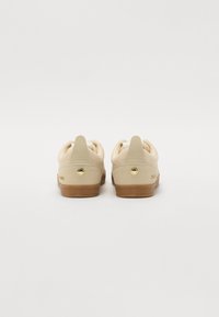 Sneaker beige con una parte superiore testurizzata, dotate di un tacco solido e dettagli in oro. La suola è in gomma marrone, progettata con un dettaglio a ritaglio rotondo.
