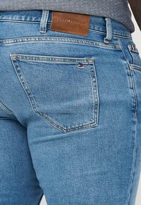 Zbliżenie na jasnoniebieskie dżinsy Tommy Hilfiger pokazujące tylną kieszeń, skórzaną naszywkę z logo marki oraz małą flagę na kieszeni i pasku.