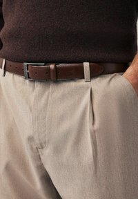 Pantalon plissé beige avec une finition texturée, associé à une ceinture en cuir marron comportant des perforations décoratives et une boucle argentée.