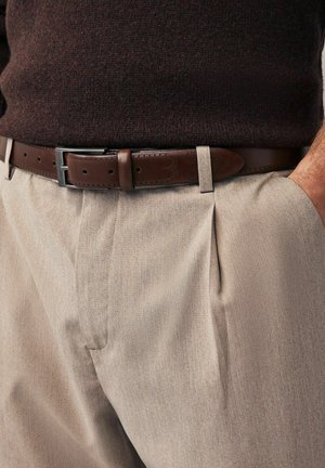 Pantalon plissé beige avec une finition texturée, associé à une ceinture en cuir marron comportant des perforations décoratives et une boucle argentée.