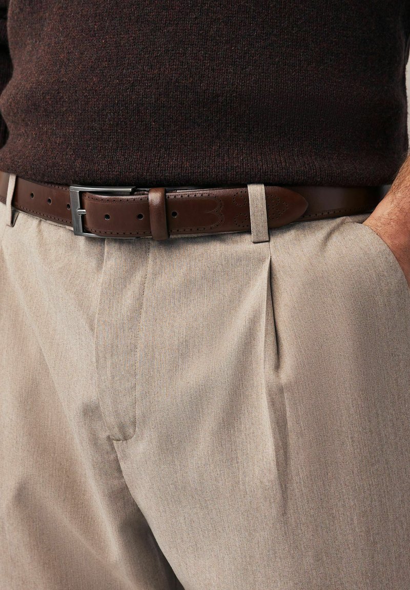 Pantalon plissé beige avec une finition texturée, associé à une ceinture en cuir marron comportant des perforations décoratives et une boucle argentée.