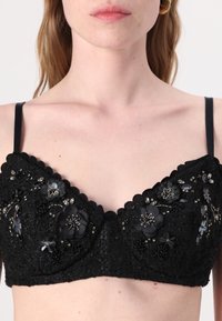 Čierny čipkovaný bralette s kvetinovou výšivkou, perličkami a akcentovými detailami. Má výrezy so zúbkami a tenké ramienka. Dekoratívne prvky sú výrazné.
