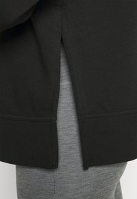 Czarna bluza z zaokrąglonym dołem i bocznymi rozcięciami, noszona na szarych, teksturowanych legginsach. Materiał wydaje się miękki i elastyczny.