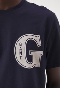 Camiseta de algodón azul marino con una gran "G" estilizada en blanco y la palabra "GANT" impresa junto a ella, con un clásico cuello redondo.