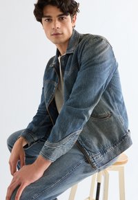 Joven con cabello rizado oscuro, vestido con una chaqueta y pantalones vaqueros azul deslavado, sentado en un taburete de madera frente a un fondo liso.