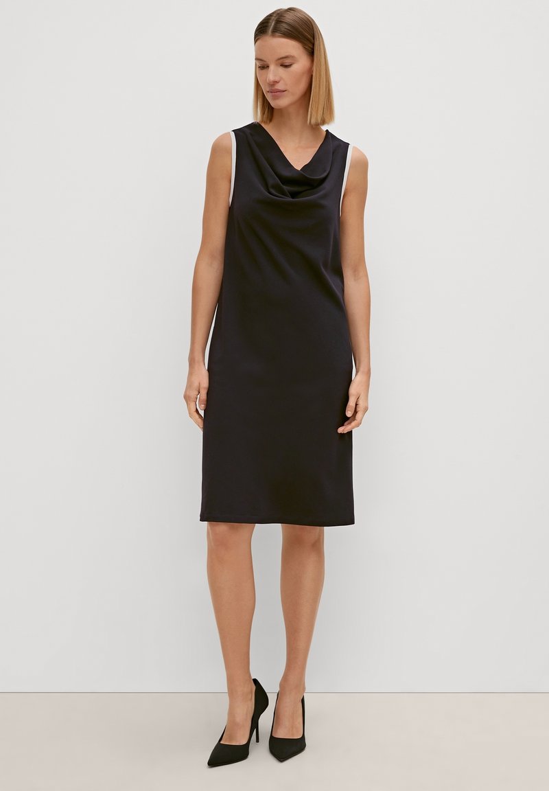 Schwarzes ärmelloses Kleid mit einem gerafften Ausschnitt, gerade geschnitten, aus glattem Stoff. Kombiniert mit schwarzen spitzen Absätzen für einen eleganten Look.