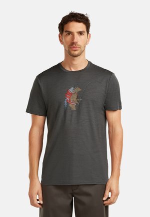 Hombre que lleva una camiseta gris oscuro con un bordado de un oso pescando, mientras lleva una mochila en el pecho.