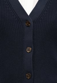 Σκούρο μπλε πλεκτό ζακέτα με τρία καφέ κουμπιά και V-λαιμόκοψη σε ανοιχτό φόντο.