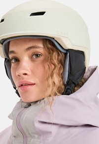 Witte skihelmet met ventilatieopeningen, logo en pluche oorkussens, gedragen door een persoon in een licht paarse jas met een hoge kraag.
