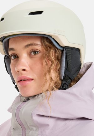Witte skihelmet met ventilatieopeningen, logo en pluche oorkussens, gedragen door een persoon in een licht paarse jas met een hoge kraag.