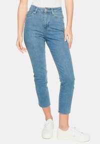 Jasnoniebieskie, wysokie, obcisłe dżinsy z postrzępionym dołem, pięcioma kieszeniami i subtelnymi detalami przeszycia; w zestawie z białymi sneakersami.