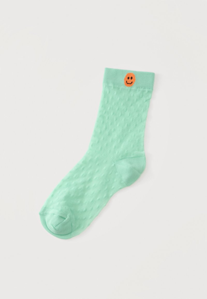 Chaussette vert clair texturée avec de petits points en relief et un visage souriant orange près du bord sur fond blanc.