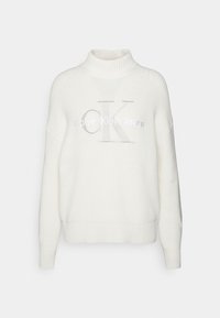 Pull en tricot blanc avec un col haut, arborant un grand logo "CK" gris et le texte "Calvin Klein Jeans" en broderie argentée.