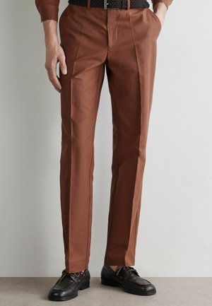 Mand iført brune, skræddersyede bukser med frontfolder, sort flettet bælte og sorte læder snørestøvler, stående mod en lys baggrund.