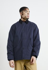Marineblauwe button-up jas met een zwarte corduroy kraag, twee zijzakken en een ontspannen pasvorm, gedragen over beige broeken.