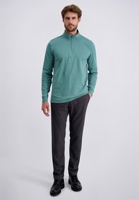 Homme debout avec une main dans la poche, portant un pull à fermeture éclair teal, un pantalon gris foncé et des chaussures de ville noires sur fond uni.