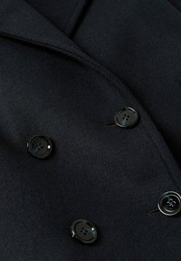 Cappotto in lana nera con design doppiopetto, rever a punte e due grandi bottoni neri con quattro fori ciascuno.