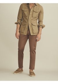 Tan-värinen utilitiopaita, jossa on läppätaskut, ruskeat slim-fit-housut ja beige espadrillot. Neutraali tausta. Luonnollinen kankaan rakenne.