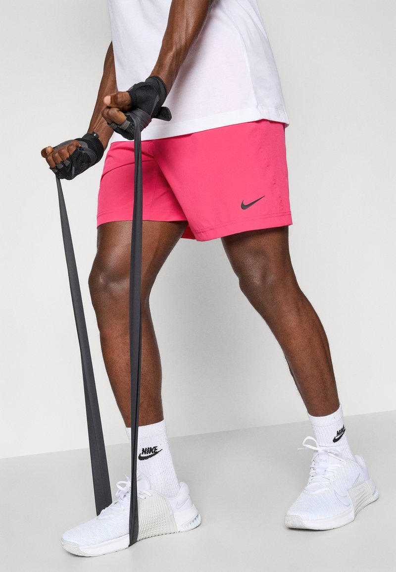 Rosa shorts med et sort Nike-logo, hvide sportsko og sorte løftehandsker, som holder sorte modstandsbånd, sat mod en neutral baggrund.