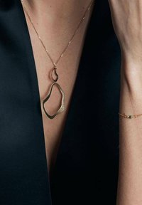 Złoty naszyjnik o płynnej formie wisi na szyi kobiety, podkreślony ciemnym strojem; na jej nadgarstku widoczna jest delikatna bransoletka.