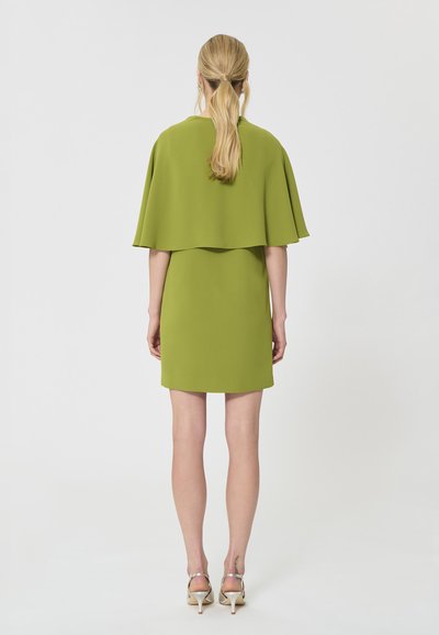 Femme avec une queue de cheval blonde portant une courte robe vert citron avec des manches cape, debout de dos, talons argentés sur un fond uni.