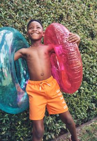 Dječak u narančastim kupaćim gaćicama drži ružičaste i plave napuhavajuće prstenove za bazen, smiješeći se na otvorenom ispred zelenog grmlja.