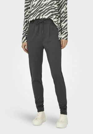 Osoba ubrana w czarne spodnie ze ściągaczem, długi rękaw w zebra print oraz białe sneakersy na platformie, stojąca na jednolitym tle.