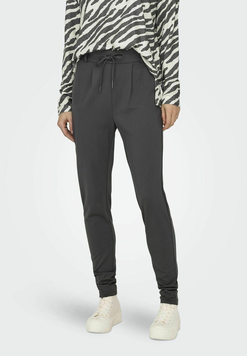 Personne portant un pantalon noir à cordon, un haut à manches longues à imprimé zèbre, et des baskets blanches à semelle compensée, debout devant un fond uni.
