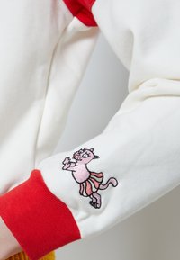 Biela mikina s červeným golierom a manžetami. Obsahuje vyšívanú ružovú karikatúru mačky v sukni, s detailným stehom na rukáve.