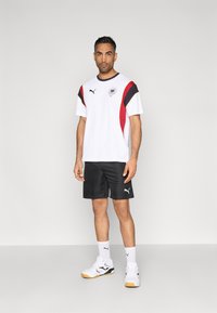 Λευκό αθλητικό μπλουζάκι με κόκκινες και μαύρες λεπτομέρειες, λογότυπο στο στήθος, συνδυασμένο με μαύρο σορτς και λευκά αθλητικά παπούτσια με μαύρες και κίτρινες λεπτομέρειες.