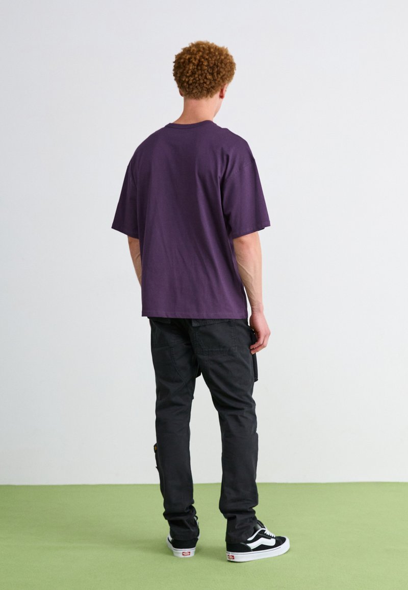 Fioletowa bawełniana koszulka, luźny krój, zestawiona z czarnymi spodniami cargo. Czarnobiałe sneakersy dopełniają strój. Prosty, casualowy design.