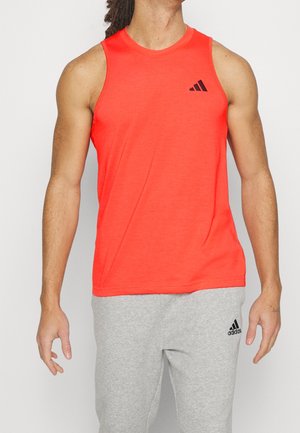 Mann trägt ein leuchtend orangefarbenes ärmelloses Sportshirt und hellgraue Adidas-Sweatpants, steht vor einem einfarbigen Hintergrund.