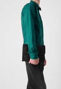 Pochette noire tenue dans une main, avec un rabat repliable et une texture lisse. Associée à une chemise à manches longues verte à boutons.