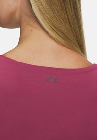 A person with long hair is shown from behind, wearing a purple top featuring a small black Under Armour logo near the neckline.